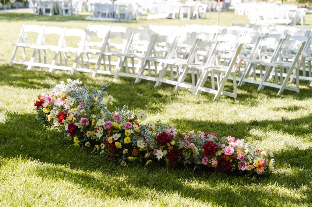 A row of colorful flowers on green grass, with white folding chairs arranged in the background for an outdoor event.