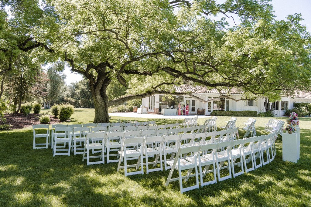 Outdoor wedding ceremony setup with white chairs arranged under a large tree, featuring a picturesque venue in the background.