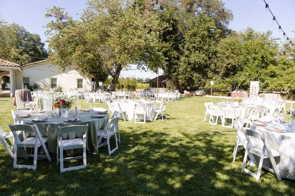 An outdoor event setting featuring multiple round tables with white chairs, elegantly arranged on a grassy area. In the background, a rustic building and trees provide a pleasant atmosphere, with overhead string lights adding to the ambiance.
