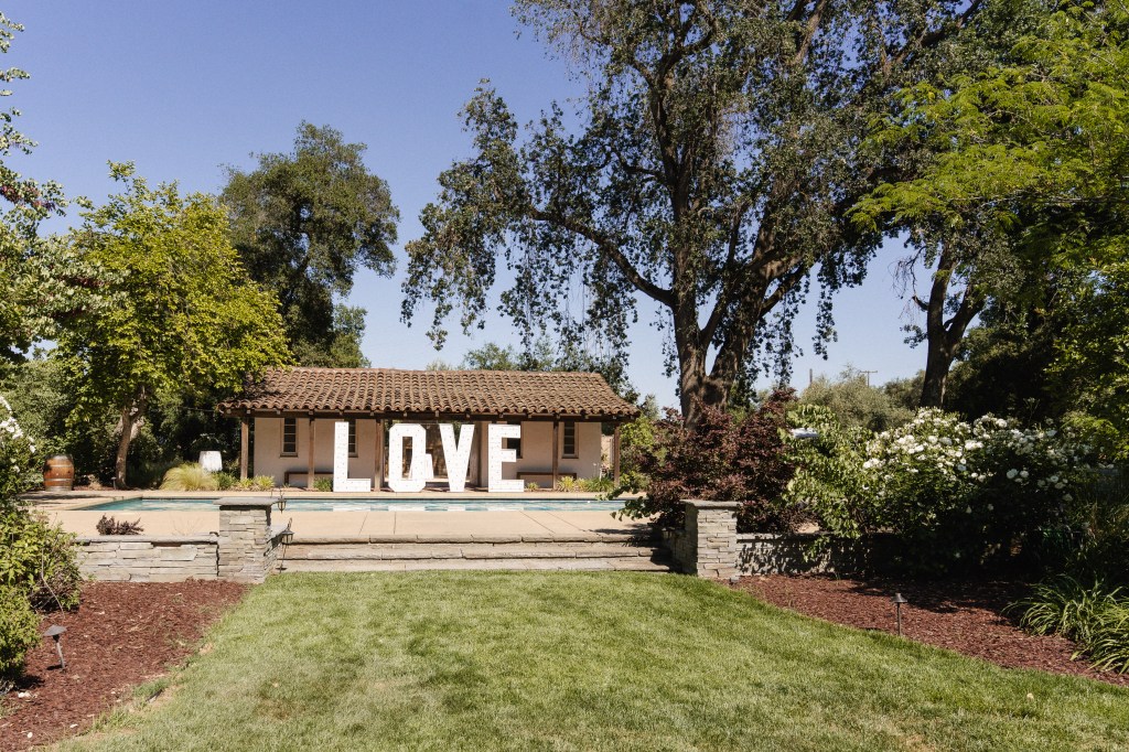 A garden view featuring a house with a brown tiled roof, framed by greenery and flowers, and large white letters spelling 'LOVE' in the foreground.