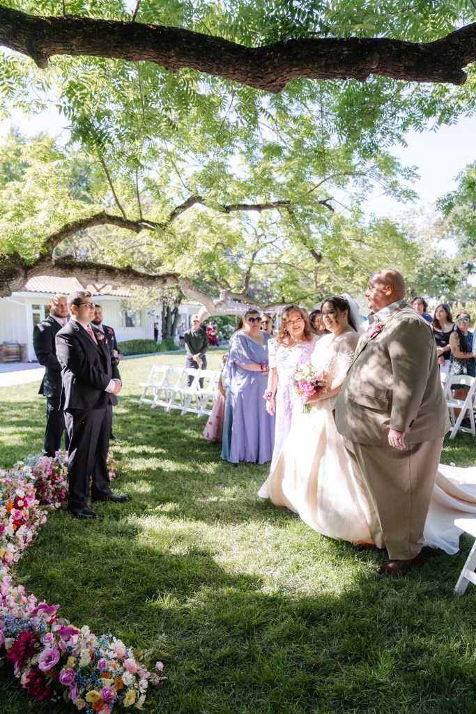 A bride, escorted by her father, walks down the aisle surrounded by guests and floral decorations under a tree.