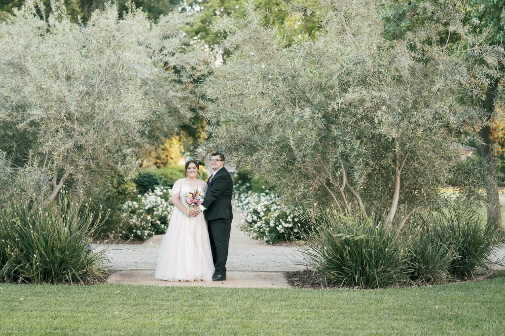 A bride in a light pink wedding dress and a groom in a black suit stand together in a landscaped garden surrounded by greenery and white flowers.