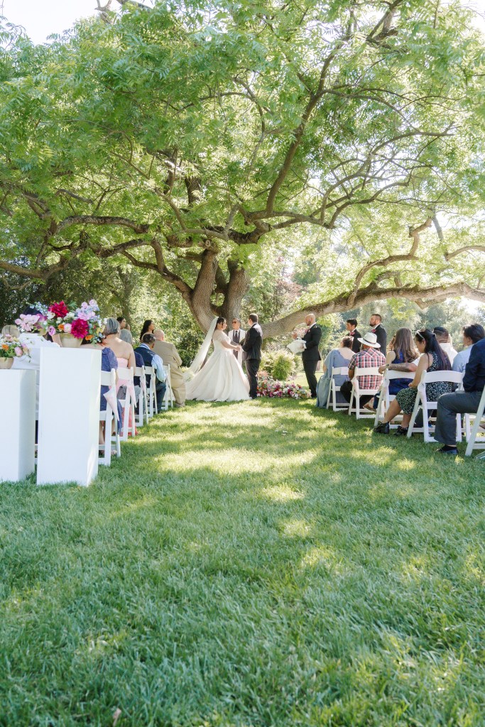 A wedding ceremony taking place outdoors under a large tree, with guests seated in white chairs. The bride and groom are standing before an officiant, surrounded by floral arrangements and lush greenery.