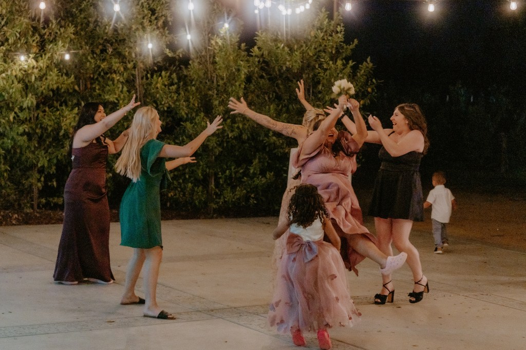 A group of women celebrating at a nighttime event, with string lights hanging above. One woman in a pink dress is joyfully catching a bouquet, surrounded by friends who are excitedly reaching out. A child in a pink dress is playfully running in the foreground.