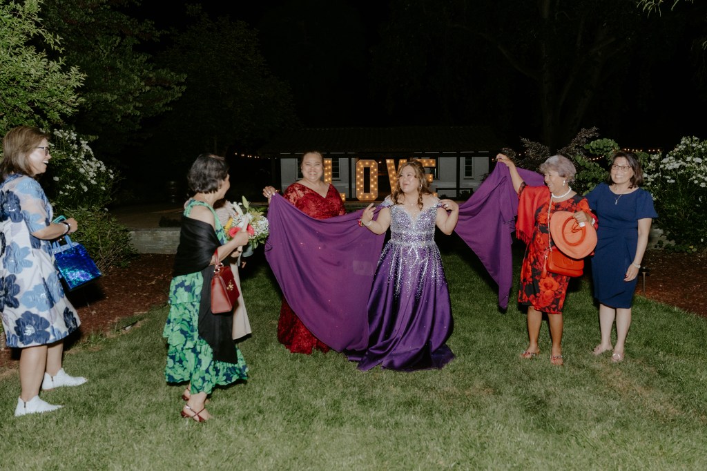 A group of women celebrating outdoors at night, with one woman in a sparkling purple gown holding a flowing purple cape, surrounded by friends dressed in colorful outfits.