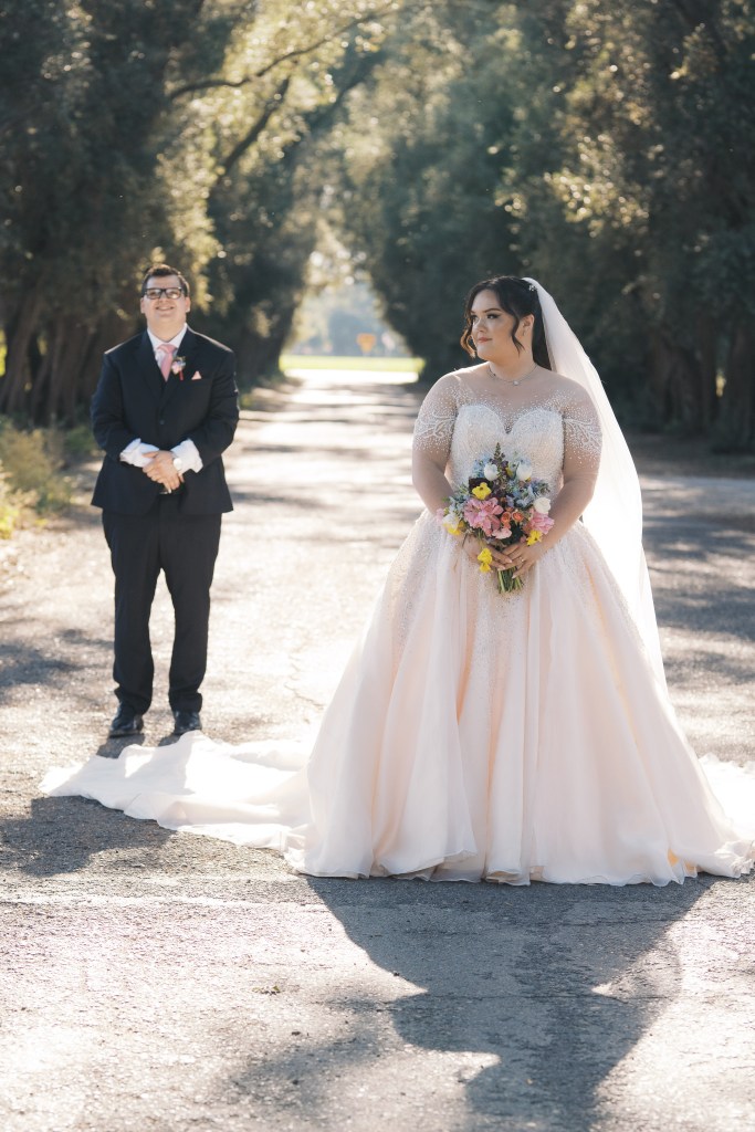 A bride in a beautiful pink and white wedding dress holds a bouquet of flowers, while a groom in a black suit stands nearby, both in a tree-lined pathway. The sunlight filters through the trees, creating a romantic atmosphere.