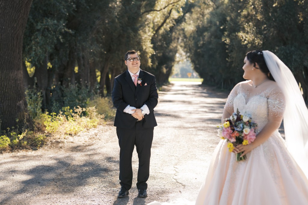 A smiling groom in a black suit stands on a gravel pathway, looking at his bride wearing a beautiful light pink wedding dress and holding a colorful bouquet. The scene is set among trees, creating a romantic atmosphere.
