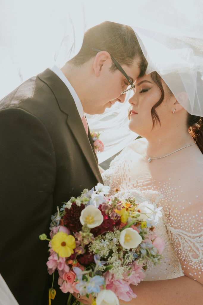 A close-up of a bride and groom sharing an intimate moment, foreheads touching, with the bride holding a colorful bouquet of flowers.