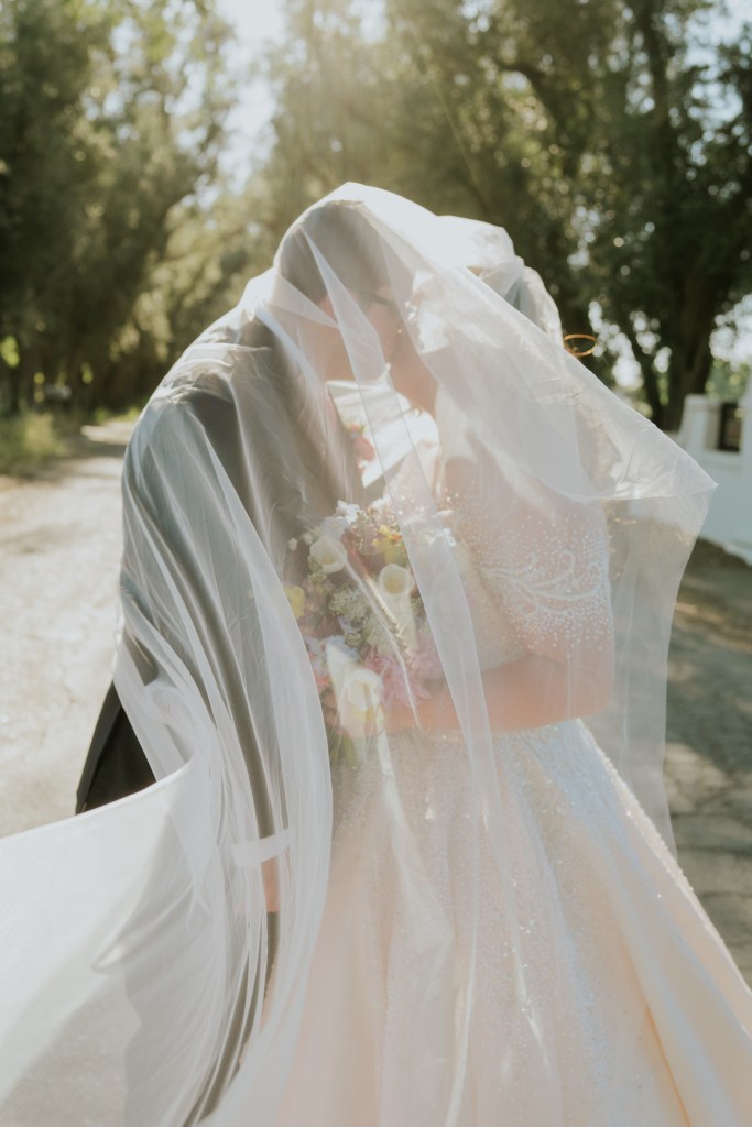 A couple sharing a romantic moment under a flowing veil outdoors, surrounded by lush trees and sunlight.