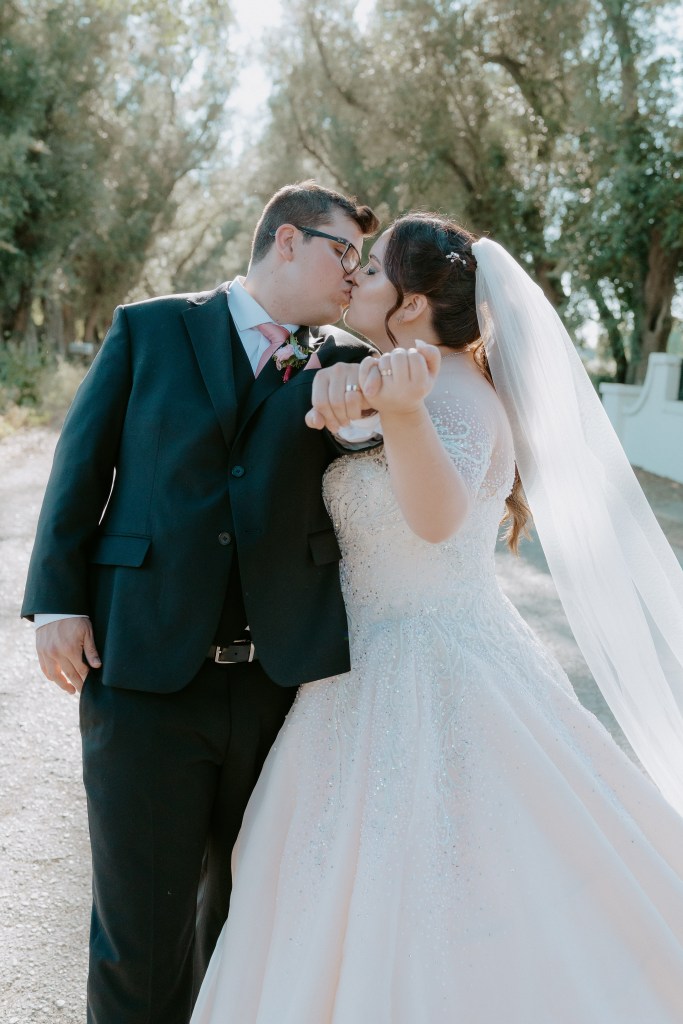 A bride and groom kiss while holding hands on a romantic outdoor path, surrounded by trees.