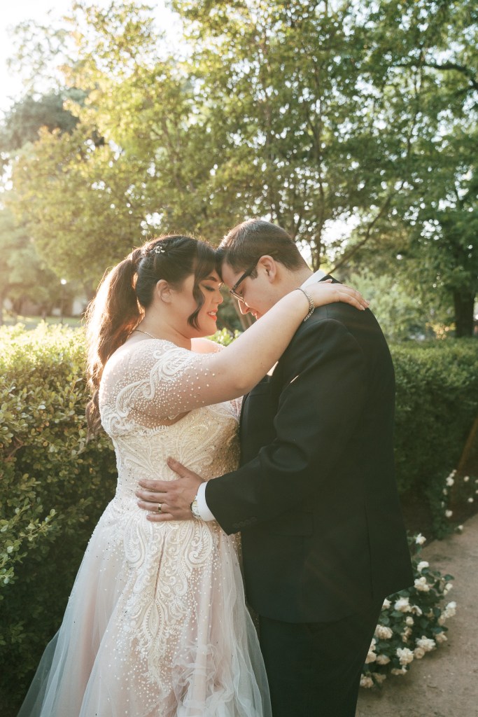 A couple in an intimate embrace, wearing a decorated wedding dress and a formal suit, surrounded by lush greenery and soft sunlight.