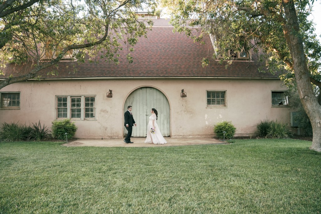 A couple in formal attire walking towards each other in front of a rustic building with a green door, surrounded by grass and trees.