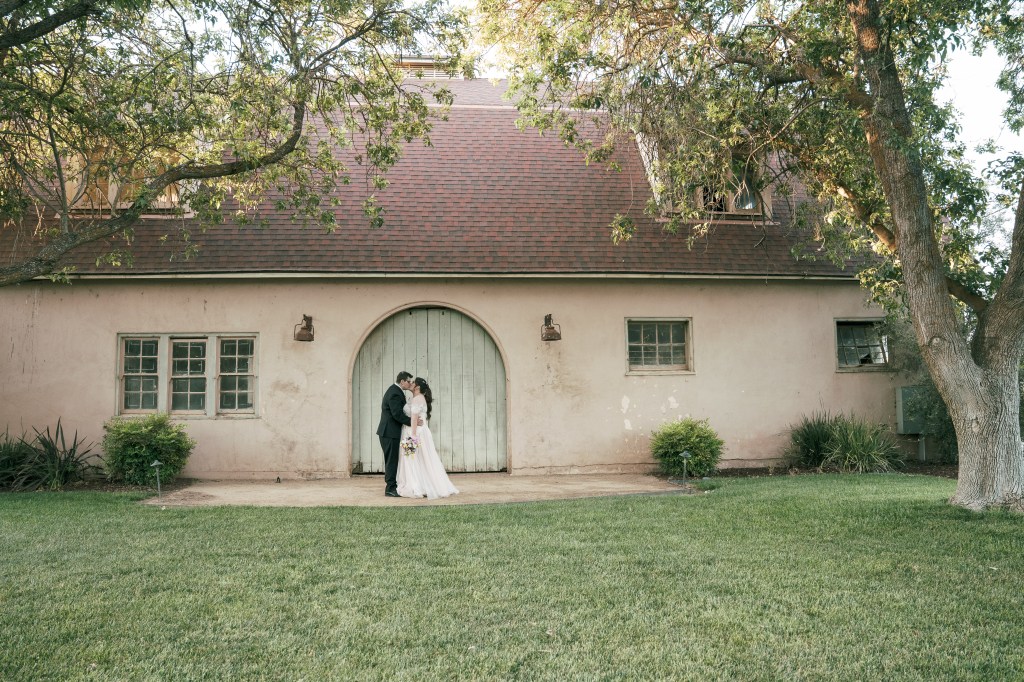 A bride and groom share a romantic kiss in front of a rustic house surrounded by greenery.