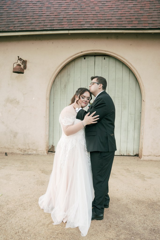A bride and groom embrace tenderly in front of a rustic building with a light green door. The bride wears a long, flowing white gown while the groom is dressed in a black suit.