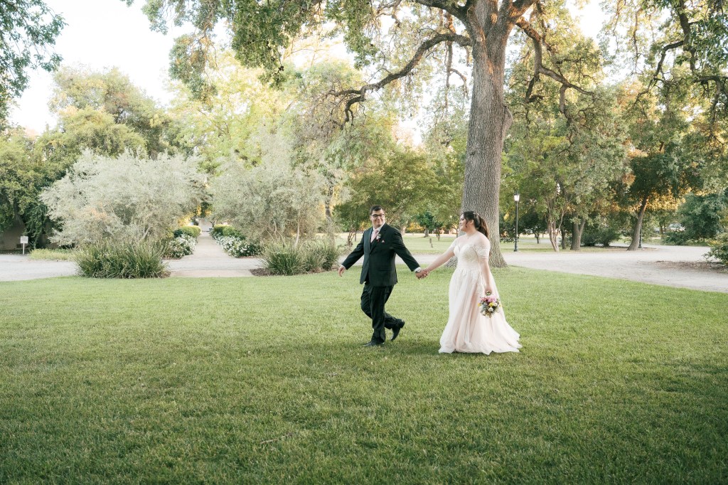 A bride and groom walking hand in hand across a lush green lawn in a park during sunset. The bride is wearing a flowing wedding dress and holding a bouquet, while the groom is dressed in a formal suit.