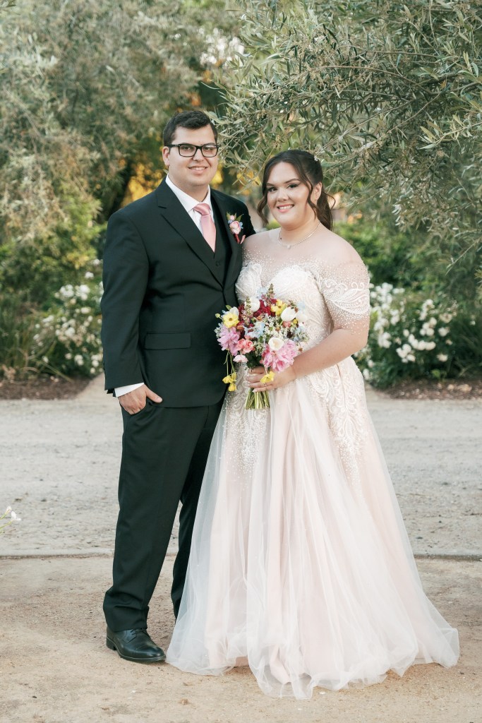 A bride and groom pose together outdoors, surrounded by greenery. The bride wears a beautiful light pink wedding gown with intricate detailing and holds a colorful bouquet, while the groom is dressed in a classic black suit with a pink tie.