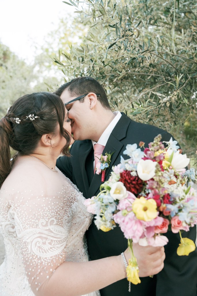 A bride and groom share a kiss outdoors, surrounded by greenery, with the bride holding a colorful bouquet.
