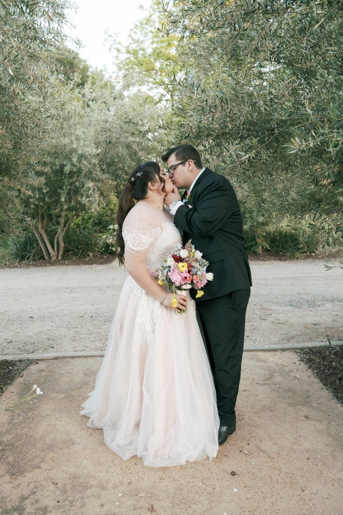 A bride and groom sharing a kiss while standing in a lush outdoor setting, surrounded by greenery. The bride is wearing a light pink wedding gown and holding a colorful bouquet of flowers.