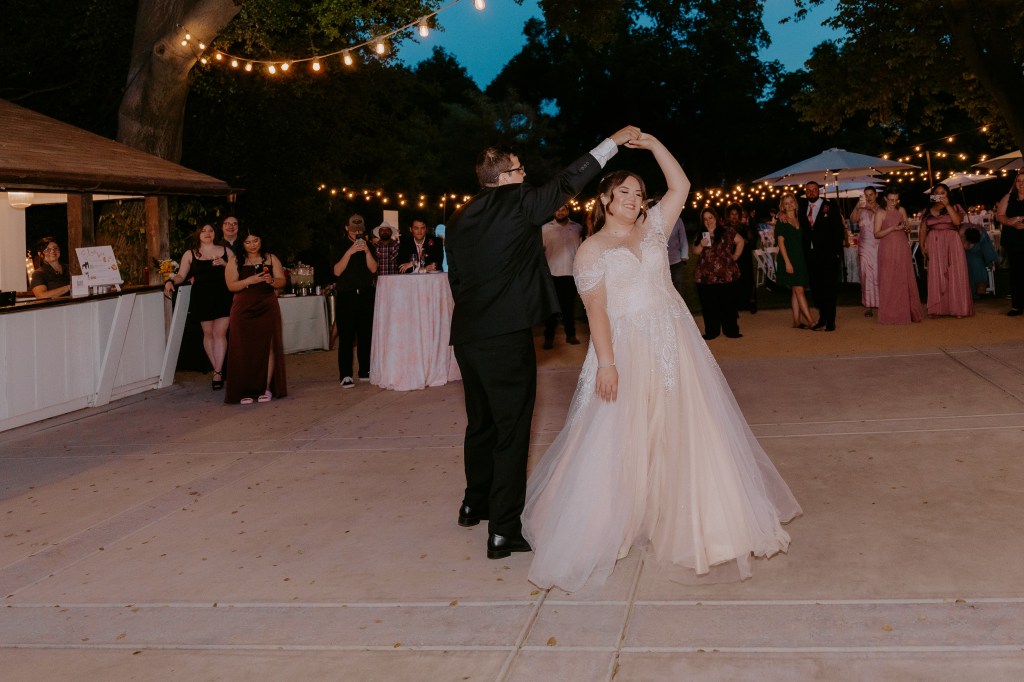 A couple dances joyfully at a wedding reception, surrounded by lively guests and string lights in the evening setting.