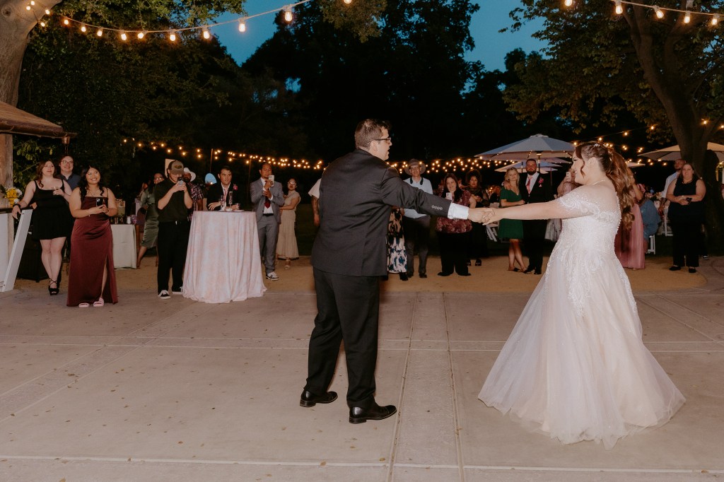 A bride and groom share their first dance at a wedding reception, surrounded by guests enjoying the celebration under string lights in an outdoor setting.