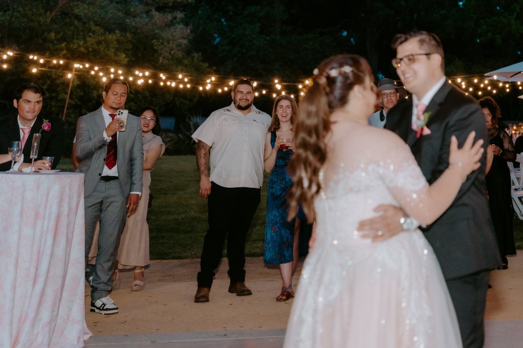 A couple dances during their wedding reception, surrounded by guests enjoying the celebration. The scene is illuminated by string lights, creating a warm and romantic atmosphere.
