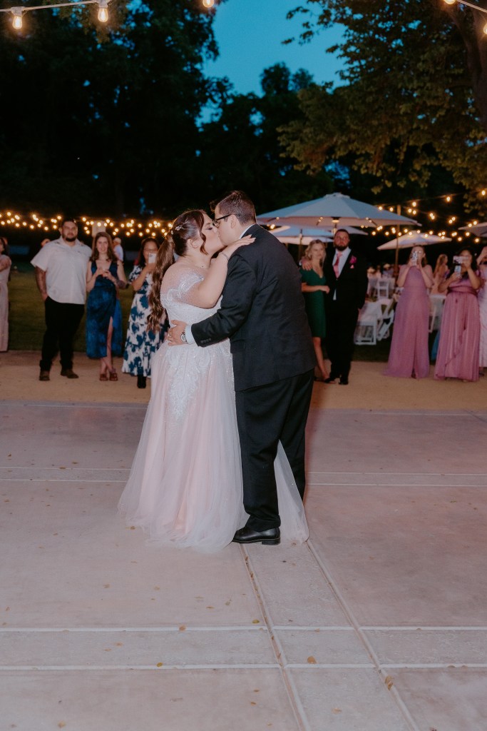 A bride and groom sharing a kiss during their wedding celebration, surrounded by guests in an outdoor setting with string lights and a twilight sky.