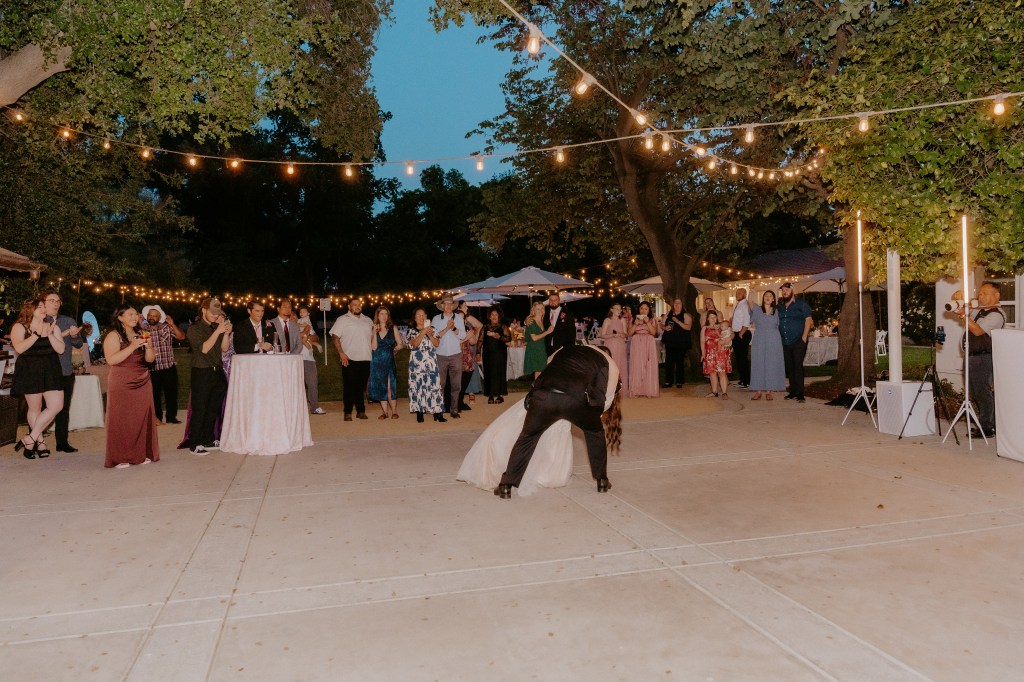 A lively outdoor event with guests enjoying themselves, including a couple dancing in the center. The scene is illuminated by string lights, with tables and various decorations visible in the background.