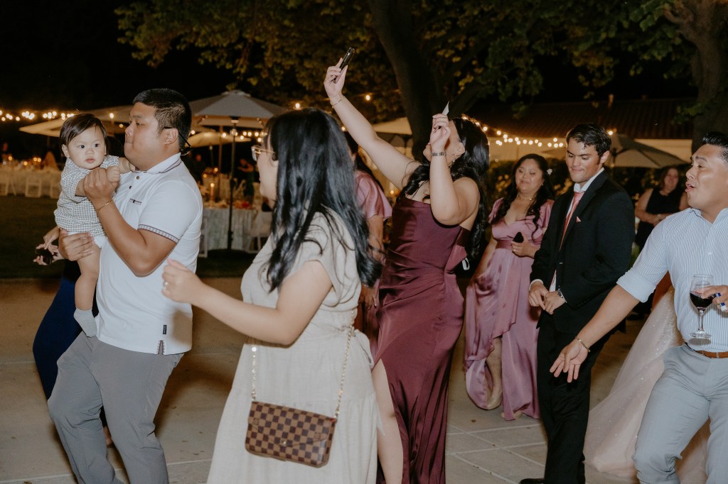 A group of people dancing at a celebration, with one person holding a baby. The atmosphere is lively with festive lighting and outdoor seating.