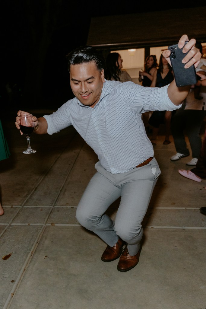 A man dancing enthusiastically at a party, holding a glass of wine in one hand and a smartphone in the other. He is wearing a striped shirt and light-colored pants.