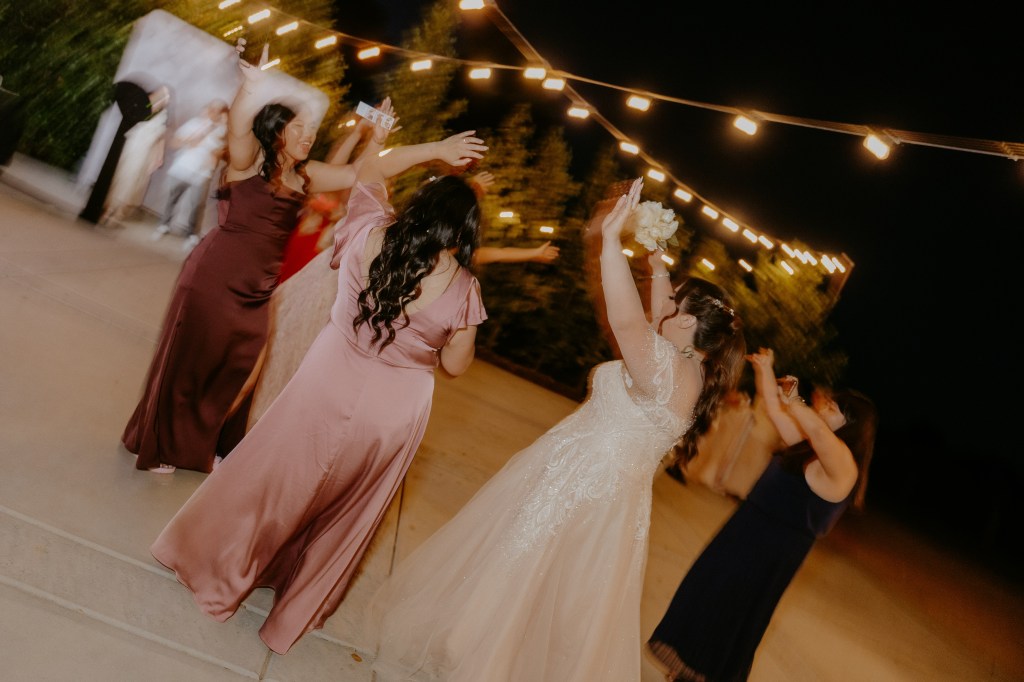 A group of women wearing elegant dresses celebrate and dance together at an outdoor evening event, illuminated by string lights.
