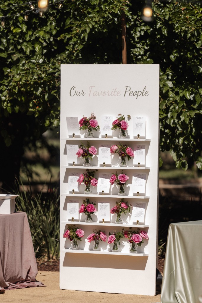 A white display board titled 'Our Favorite People' featuring several small floral arrangements with pink roses and accompanying cards, set in an outdoor environment.