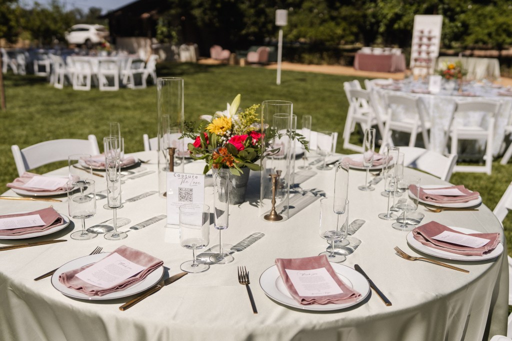 A beautifully set dining table in an outdoor setting, featuring floral centerpieces, elegant plates, folded napkins, and glassware, with additional tables visible in the background.