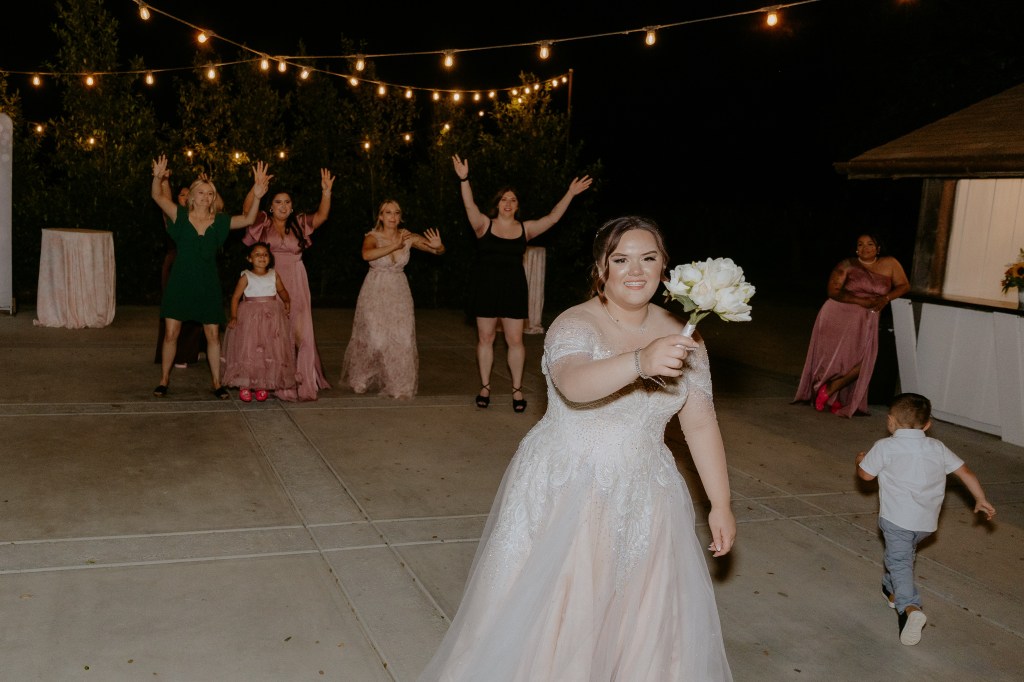 A bride in a wedding dress holds a bouquet, smiling and tossing it towards a group of excited women and children celebrating at night under string lights.