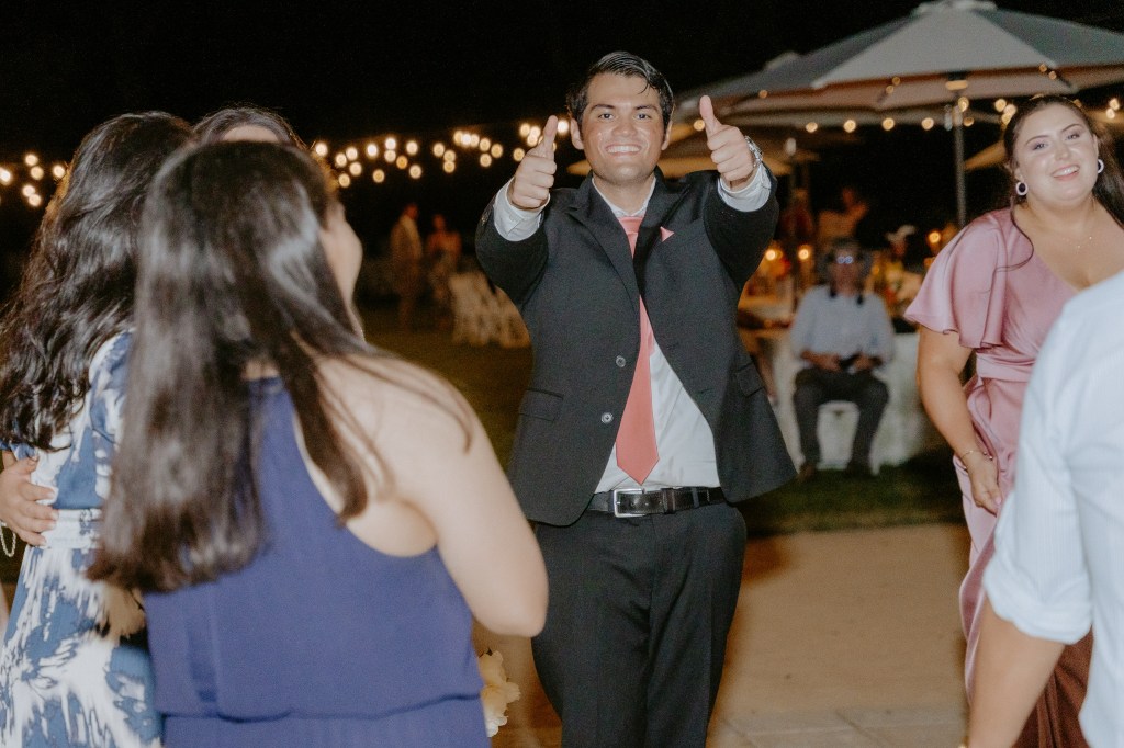 A man in a suit gives a thumbs up while celebrating at a festive outdoor event, surrounded by people dancing and string lights in the background.