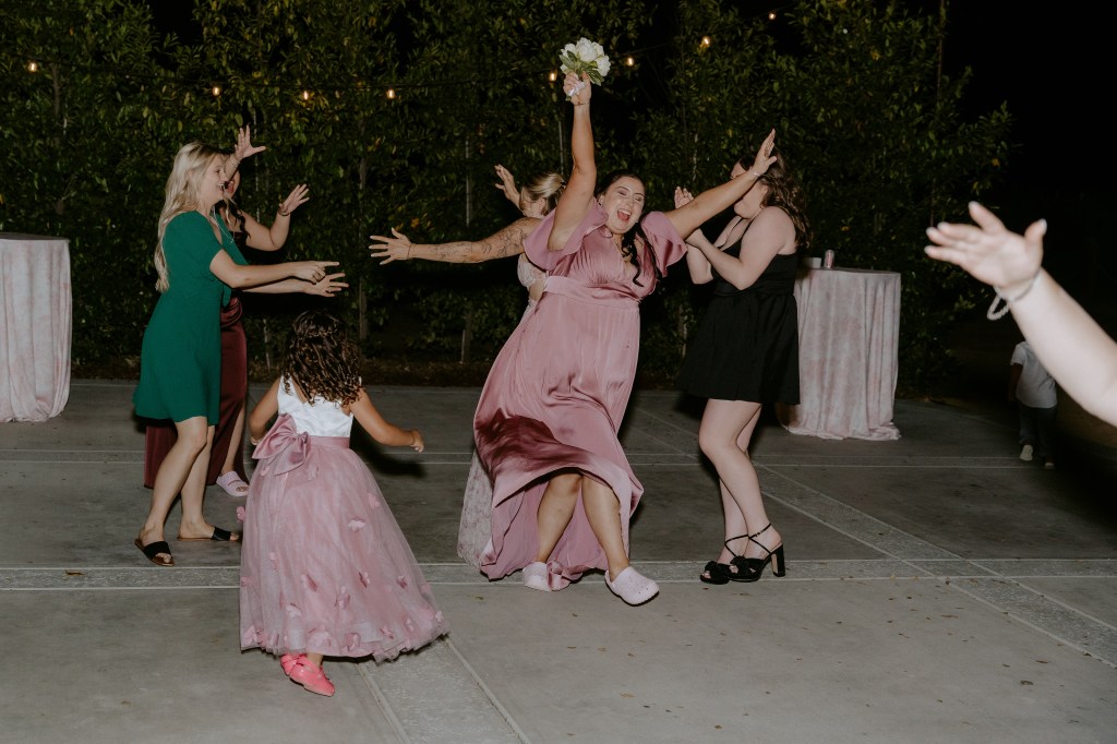 A joyful group of women celebrating and dancing at an evening event, with one woman in a pink dress playfully jumping and holding a bouquet. Children are also present, adding to the festive atmosphere.