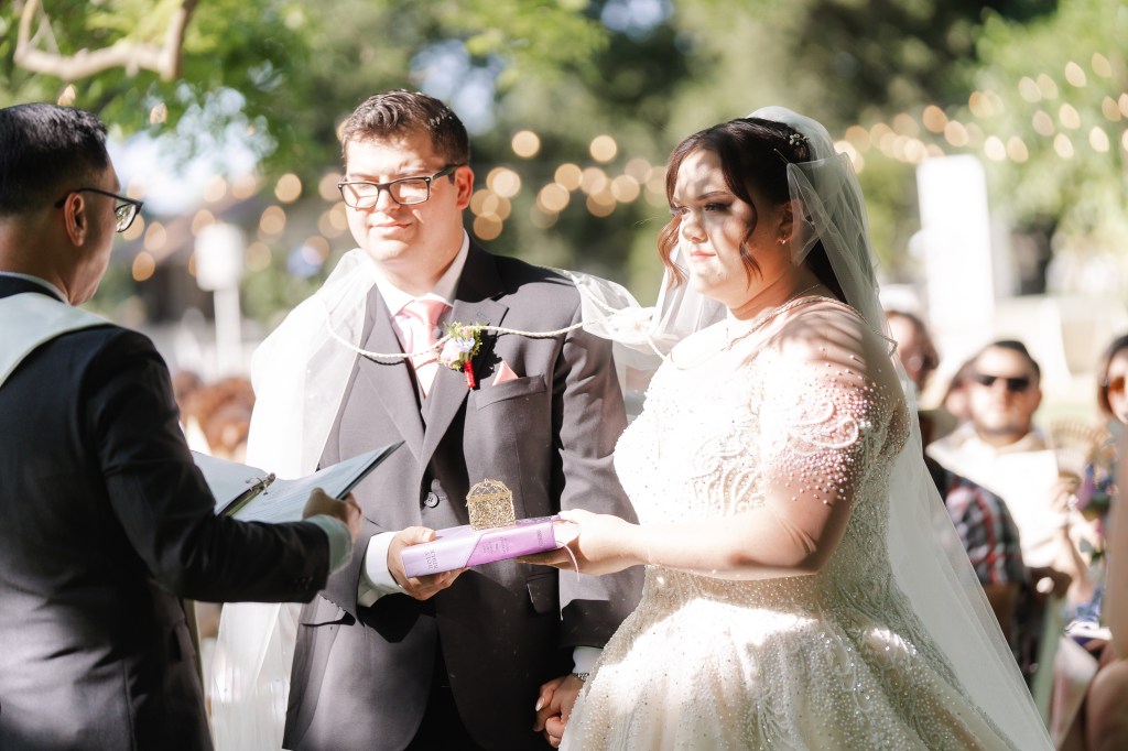 A bride and groom standing together during their wedding ceremony, with the groom holding a book and the bride looking at him. The scene is set outdoors with soft lighting and guests in the background.
