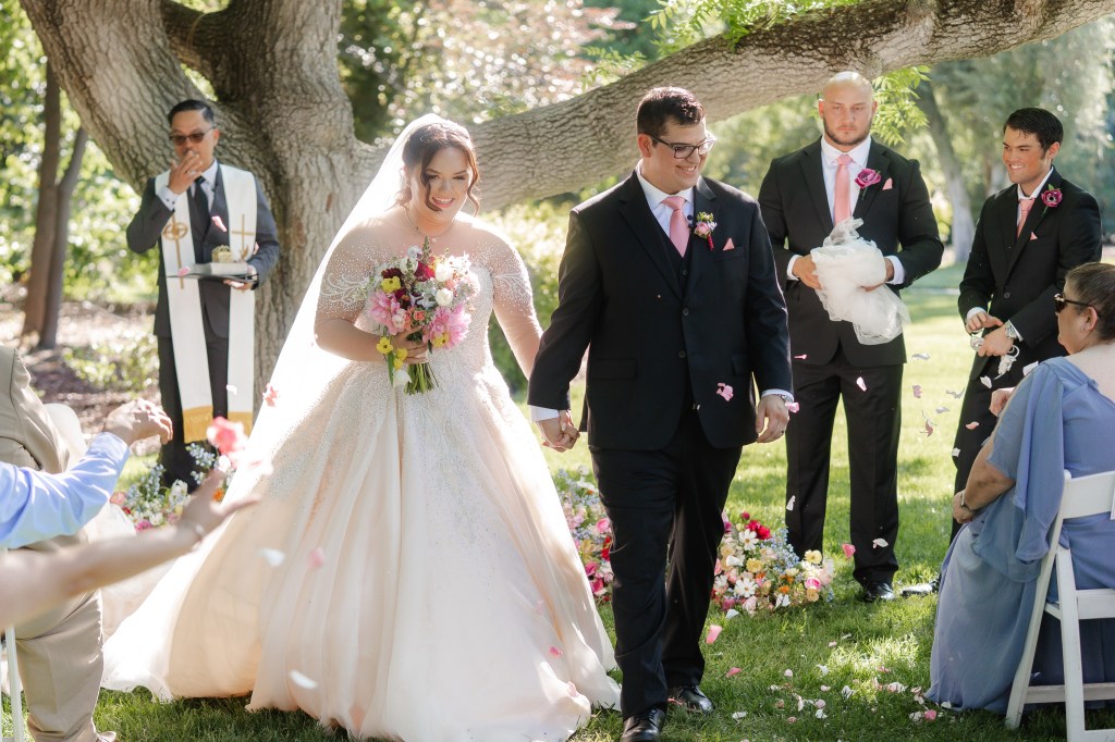 A joyful bride and groom walk hand in hand after their wedding ceremony, surrounded by guests, with petals being thrown in celebration. The bride is in a sparkling, light pink gown, holding a colorful bouquet, while the groom is dressed in a black suit with a pink tie. In the background, a minister stands beneath a tree.