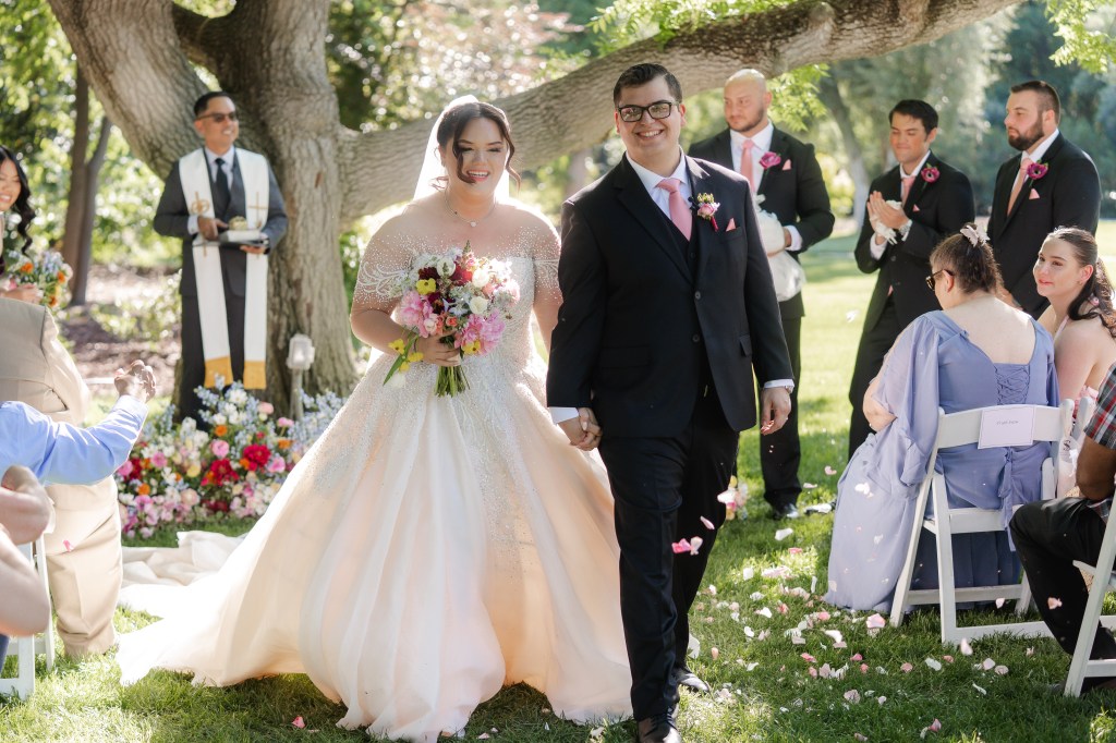 A newlywed couple walks hand in hand down the aisle during their outdoor wedding ceremony, surrounded by guests and floral arrangements.